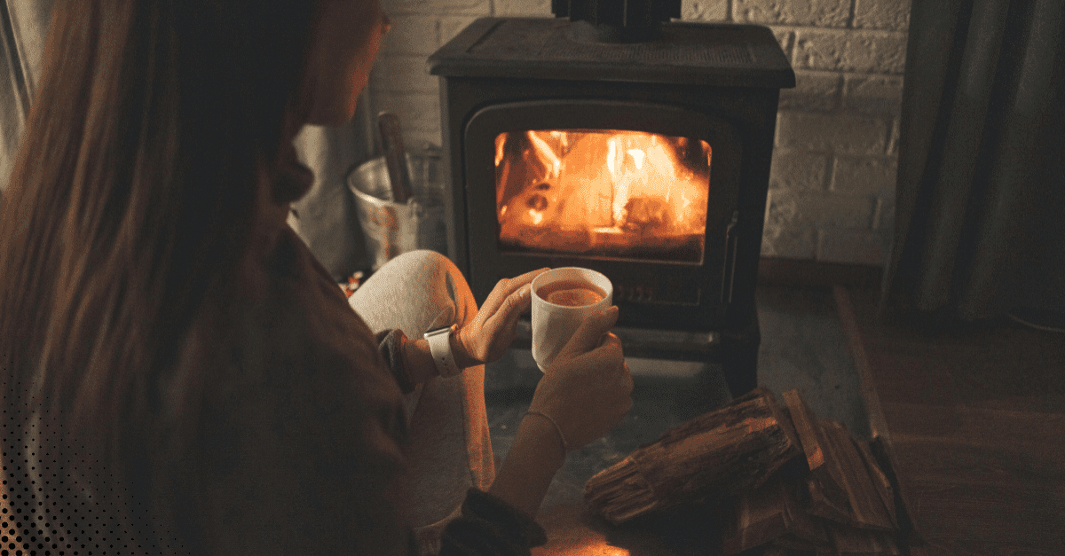 A person sitting on the floor, enjoying a hot drink in front of a wood-burning stove with a cozy fire.